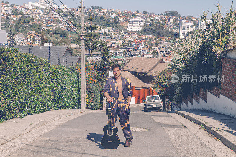 在Viña del Mar，瓦尔帕莱索，智利，一名肌肉发达的时尚男子，穿着蓝色西装外套，手拿原声吉他站在街道中央。时尚的年轻人。